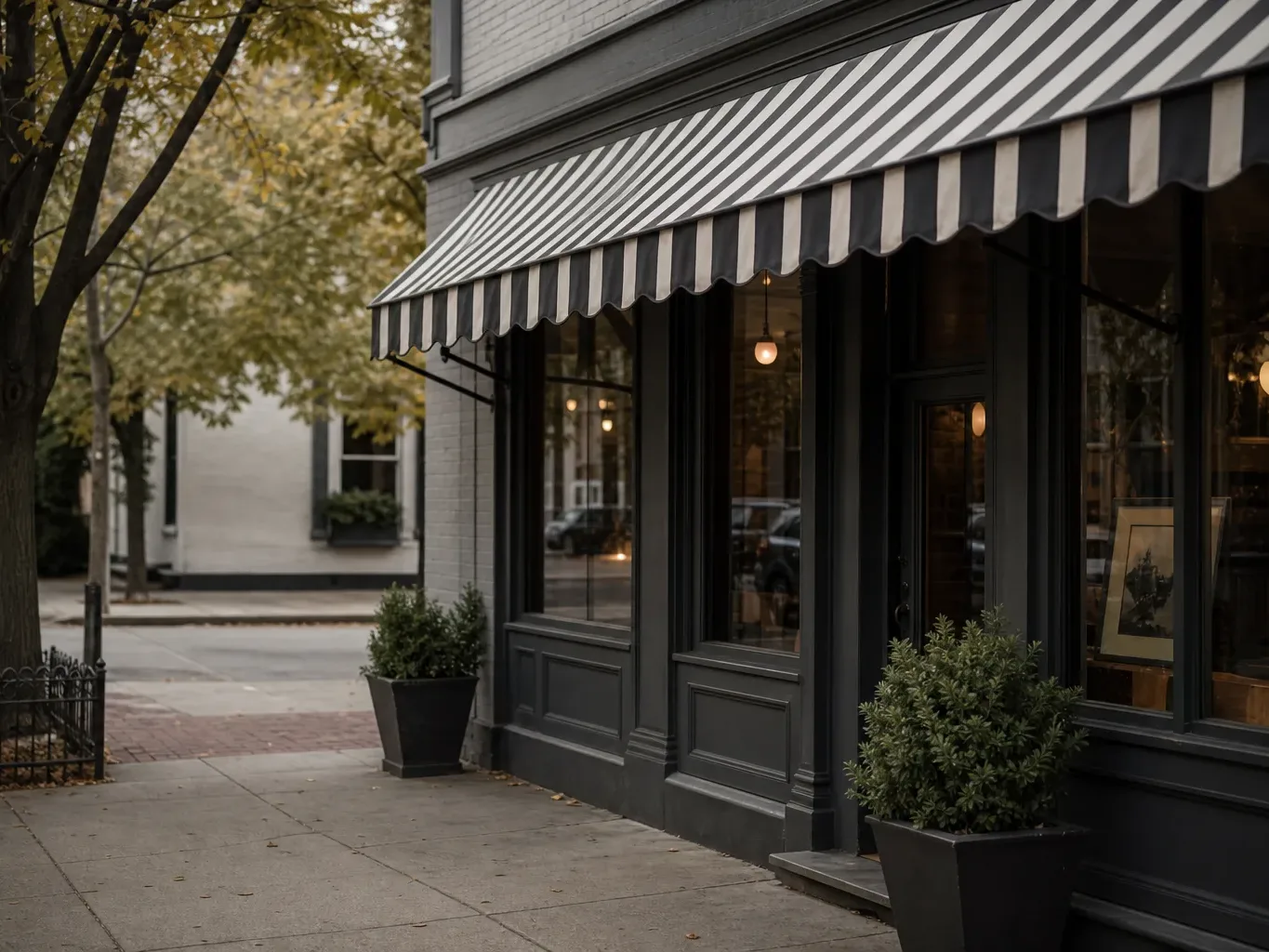 A dark-painted shopfront with a black-and-white striped awning, large windows with warm interior lighting, potted box shrubs, and autumn trees lining the empty pavement