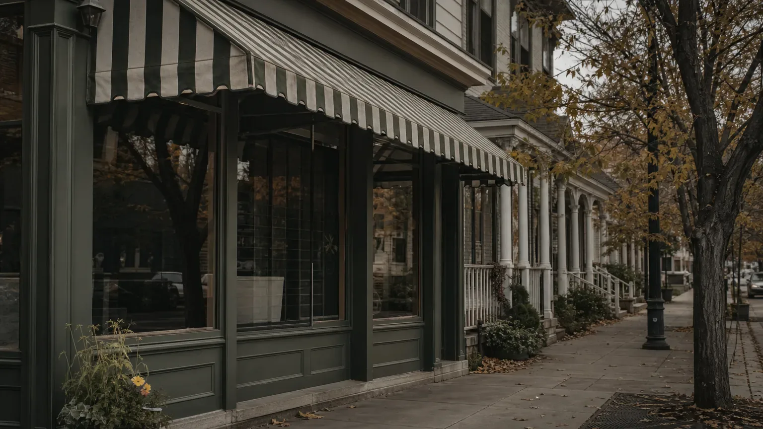 A row of dark green shopfronts with green and white striped awnings lines a quiet pavement, with trees in autumn colour and wooden porches visible further along the block