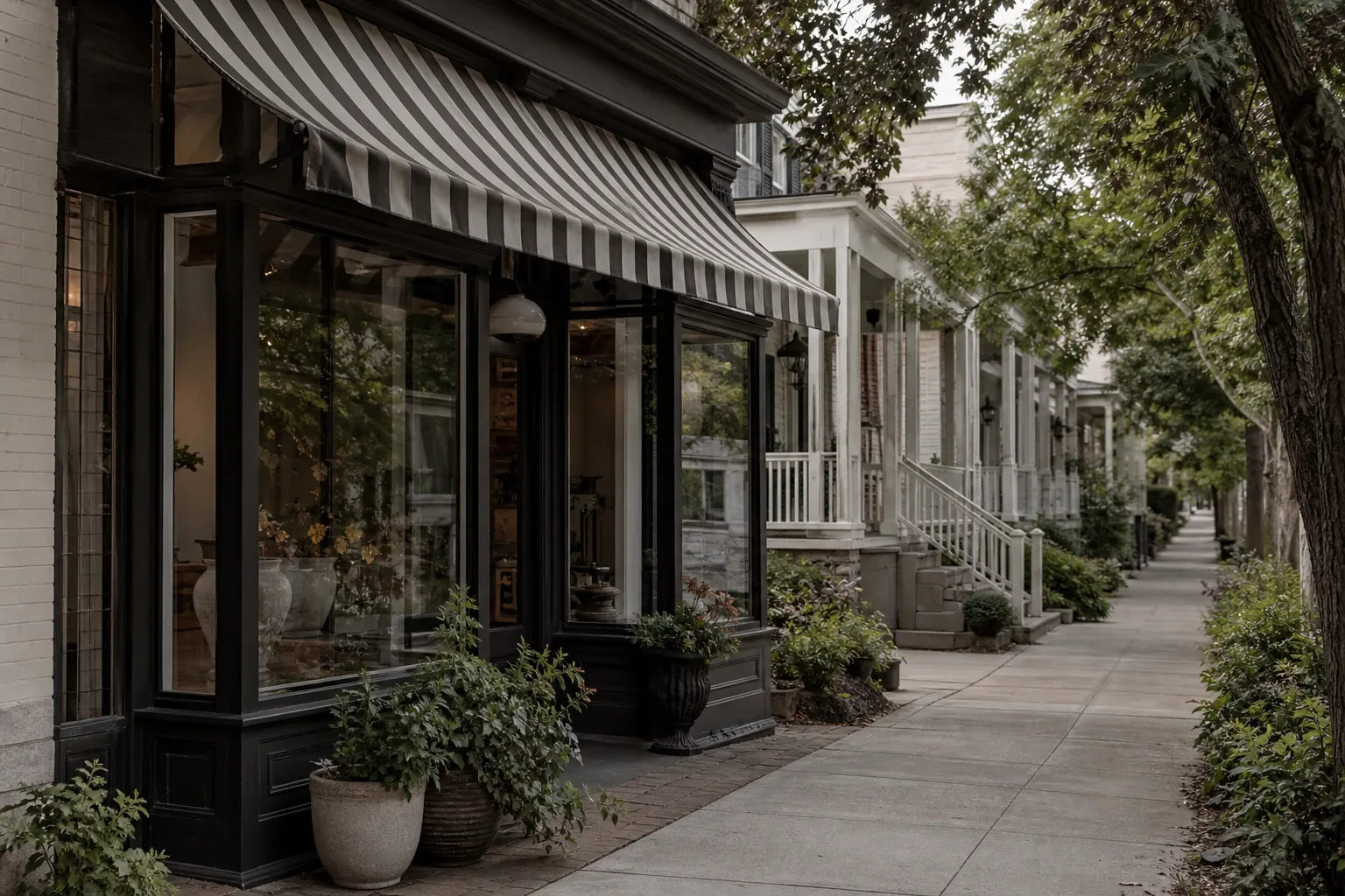 A shopfront with a black-framed bay window and grey-and-white striped awning on a tree-lined pavement, with white-porched residential buildings receding into the distance
