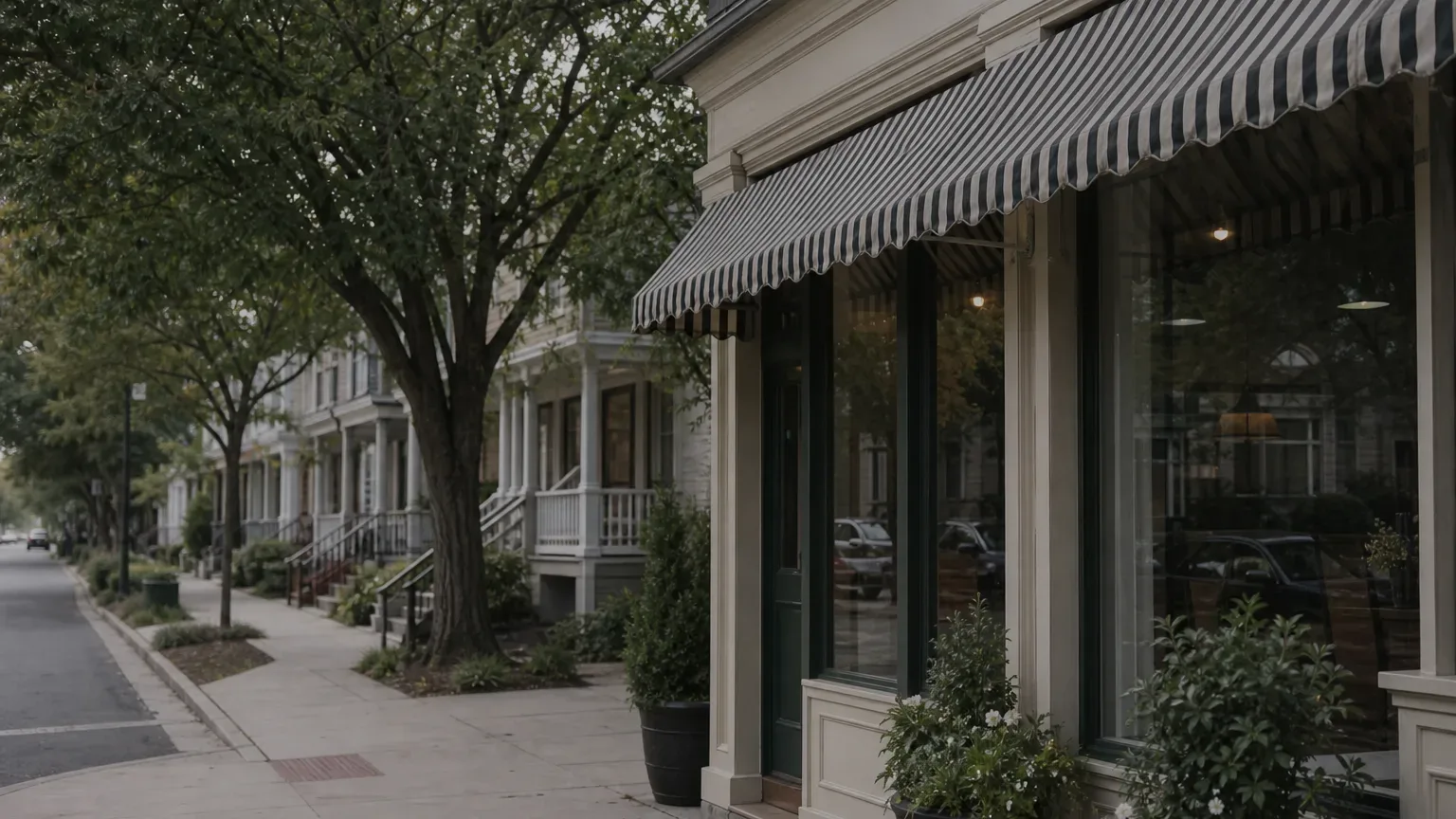 A shopfront with striped awnings and potted plants lines a quiet pavement beside mature trees, with wooden-porched buildings stretching into the distance