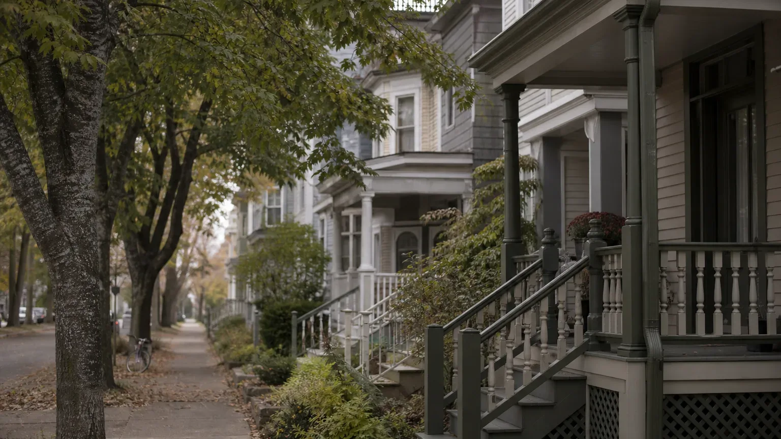 A tree-lined residential pavement stretches past a row of wooden-porched houses with painted railings and staircases in soft autumn light