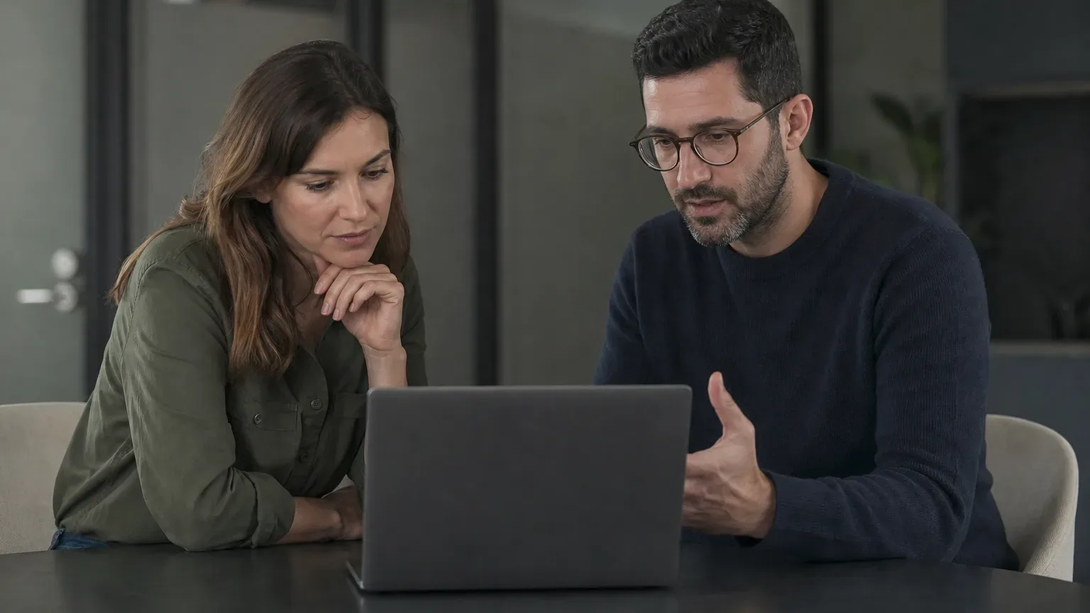 Two people sit side by side at a dark desk in an office, both looking at a laptop, one gesturing towards the screen whilst the other listens attentively
